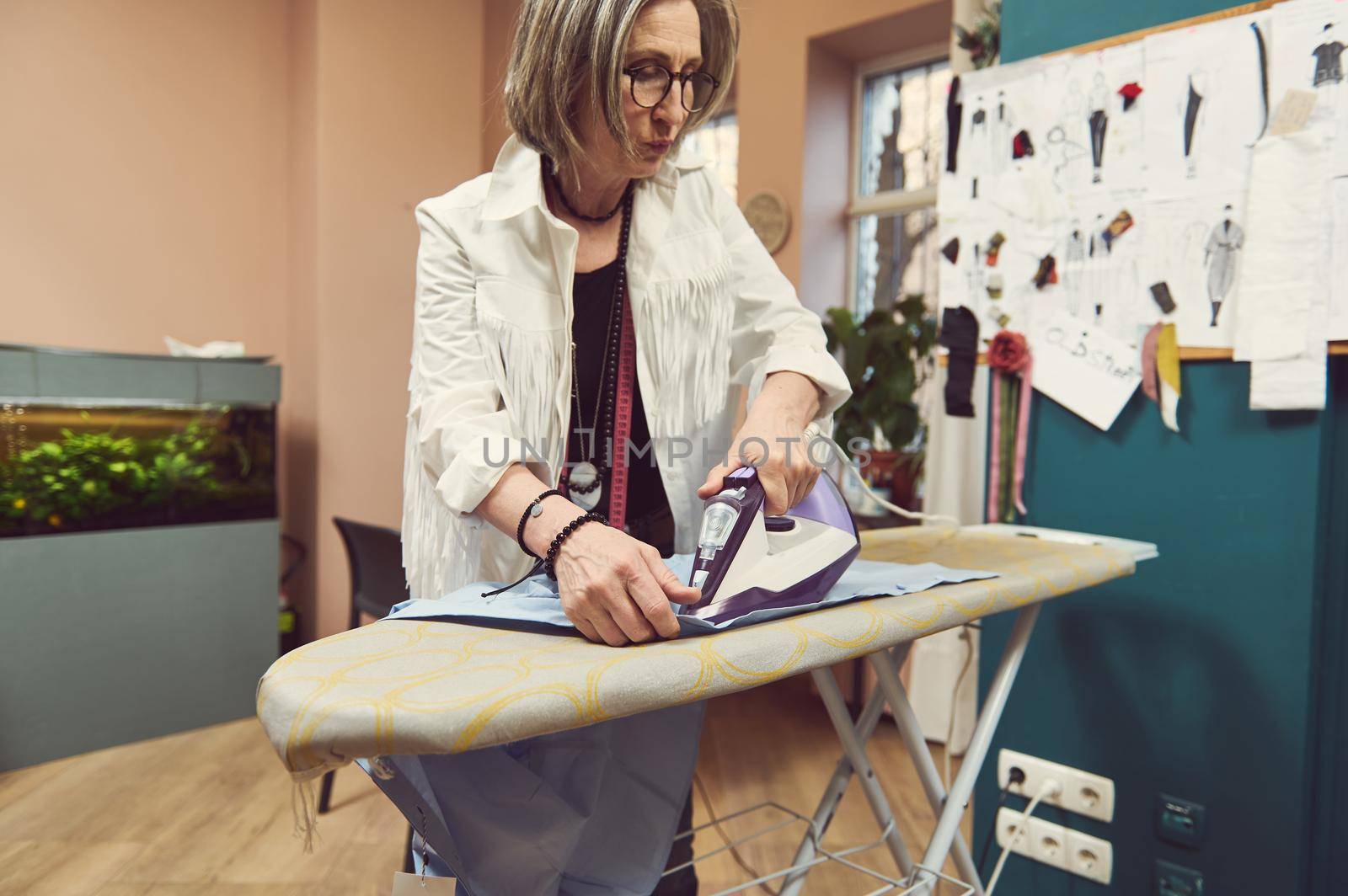 Fashion designer tailor using steam iron to press blue shirt from new collection on an ironing board in a clothing design and tailoring atelier by artgf