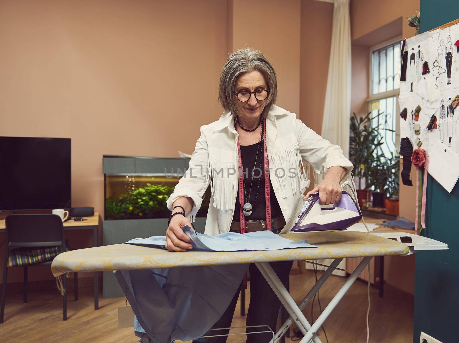 Mature white-haired Caucasian woman seamstress, tailor ironing blue shirt with steaming on the iron board before trying in on a mannequin in fashion design atelier by artgf