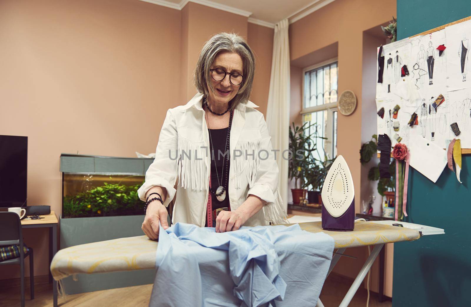 Mature 60 years old European woman fashion designer tailor ironing blue shirt from new collection in a tailoring atelier. Seamstress, sewer, craftswoman, needlewoman, dressmaker, fashion stylist by artgf