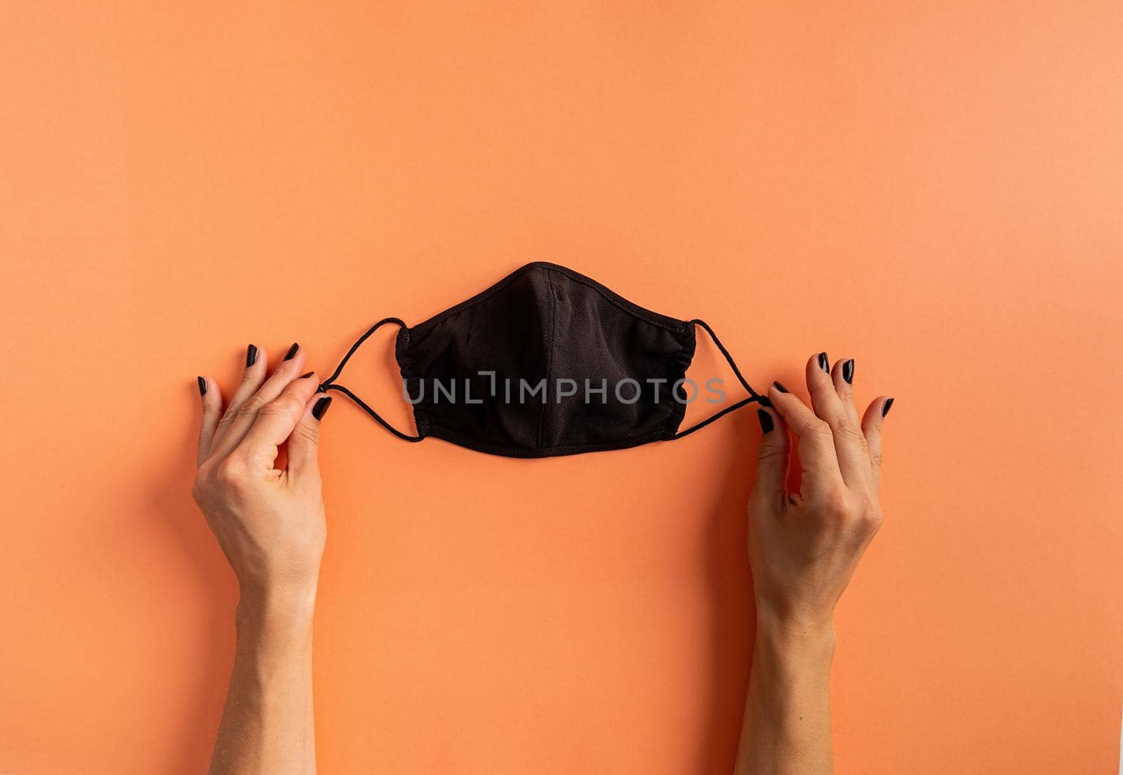 Happy Halloween concept. Social distancing and virus protection. Woman hand with black nails holding black protective mask on orange nackground