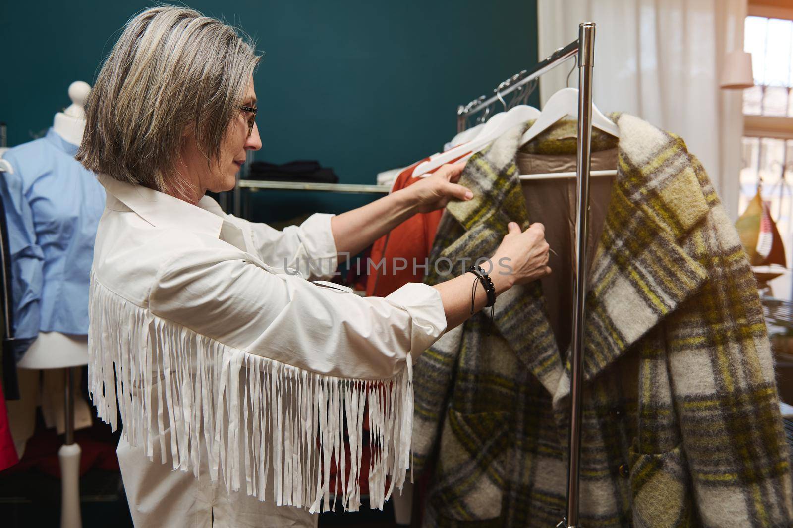 Caucasian mature woman, trendy fashion designer, tailor, seamstress standing in a garment storeroom, looking through the hangers with clothes ready for alteration. Tailoring, fashion design concept