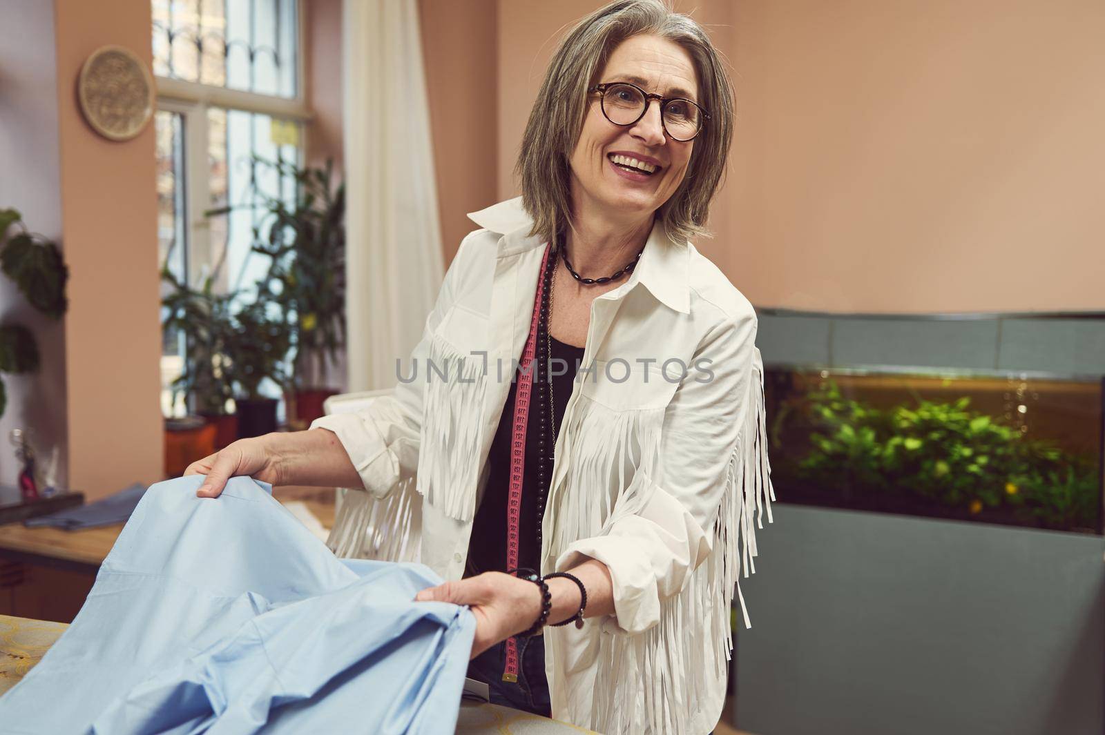 Positive elderly gray-haired Caucasian woman ironing fabric, preparing for sewing, working in a fashion design and repair atelier. Fashion designer, seamstress, dressmaker, tailor workplace