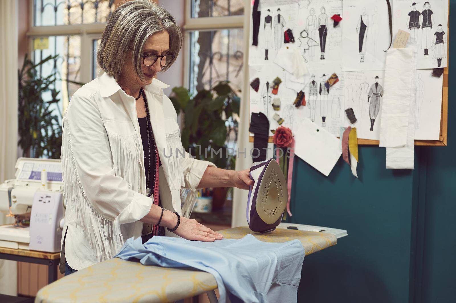 Positive elderly gray-haired European woman ironing shirt, working in a fashion design and repair atelier. Fashion designer, seamstress, dressmaker, tailor workplace by artgf
