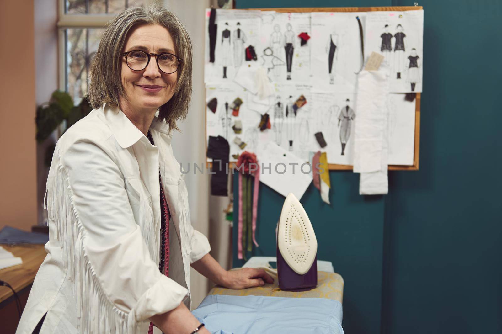 Charming stylish elderly European gray-haired woman smiles looking at camera, standing at ironing board and stroking fabric with steaming iron in her fashion clothing design and repair atelier