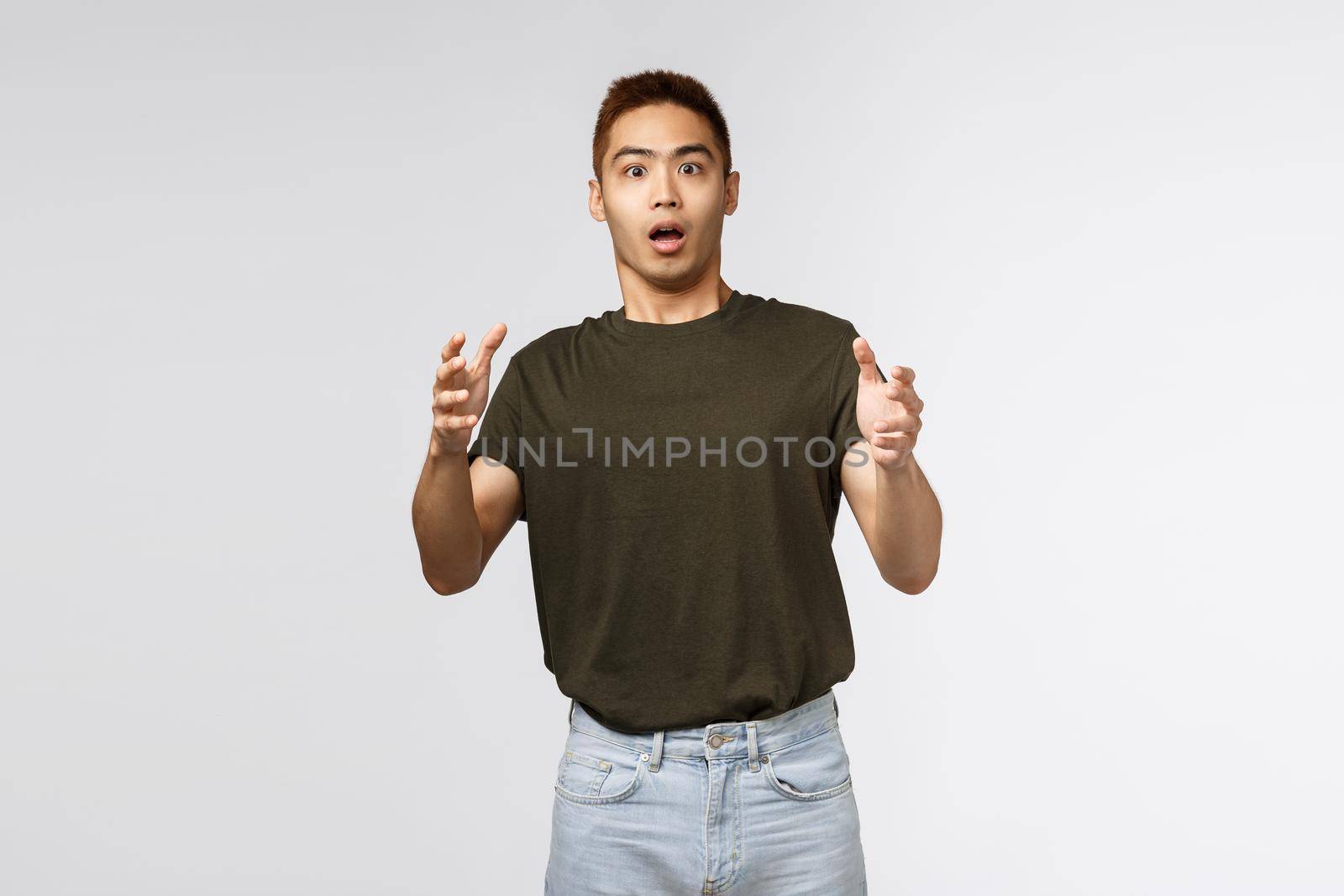 Portrait of shocked young male, taiwanese guy catching product that person throwing at him, raising hands up and look intense and stressed, gasping startled, standing grey background.