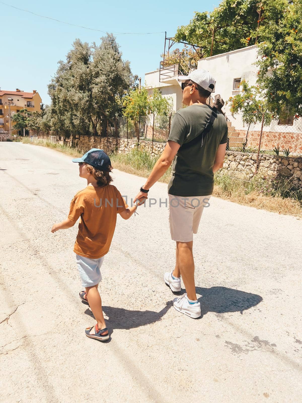 A man father dad holding his child kid school boy hand while taking a walk stroll outside on a sunny hot day by Ostanina