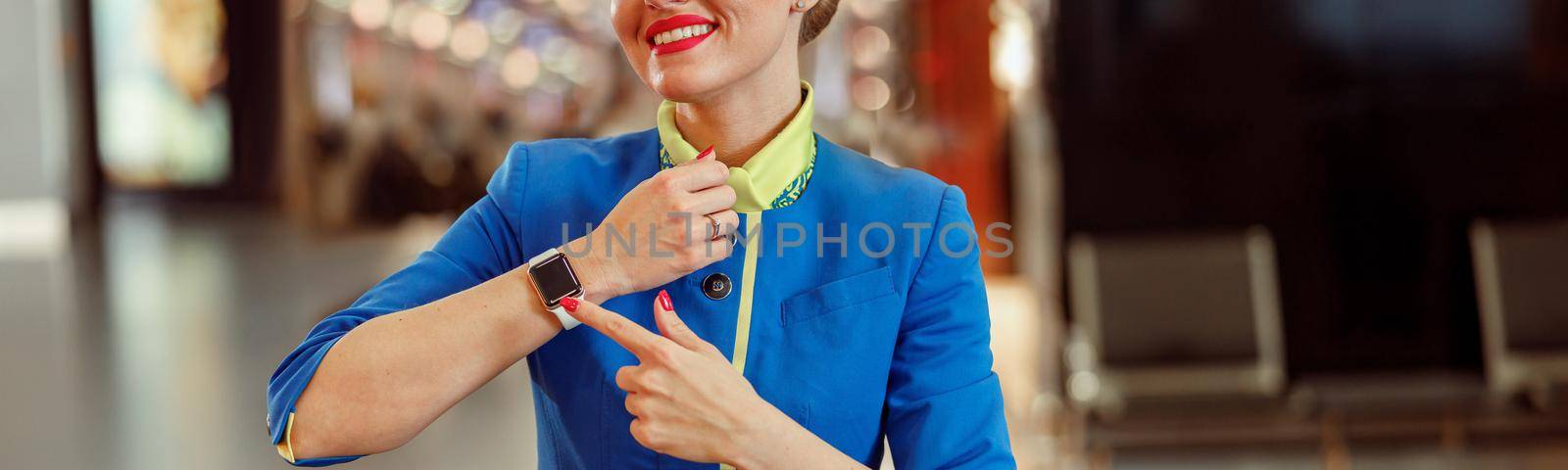 Joyful female flight attendant in air hostess uniform showing her wristwatch and smiling while standing in airport terminal