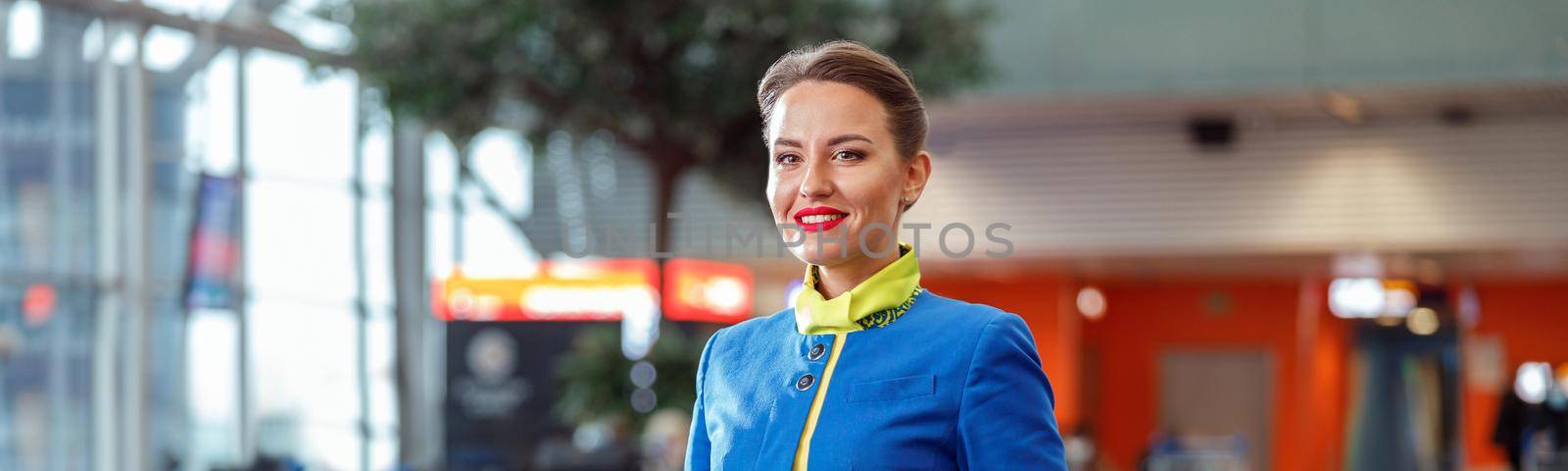 Cheerful female flight attendant in airline air hostess uniform looking at camera and smiling while standing in passenger departure terminal