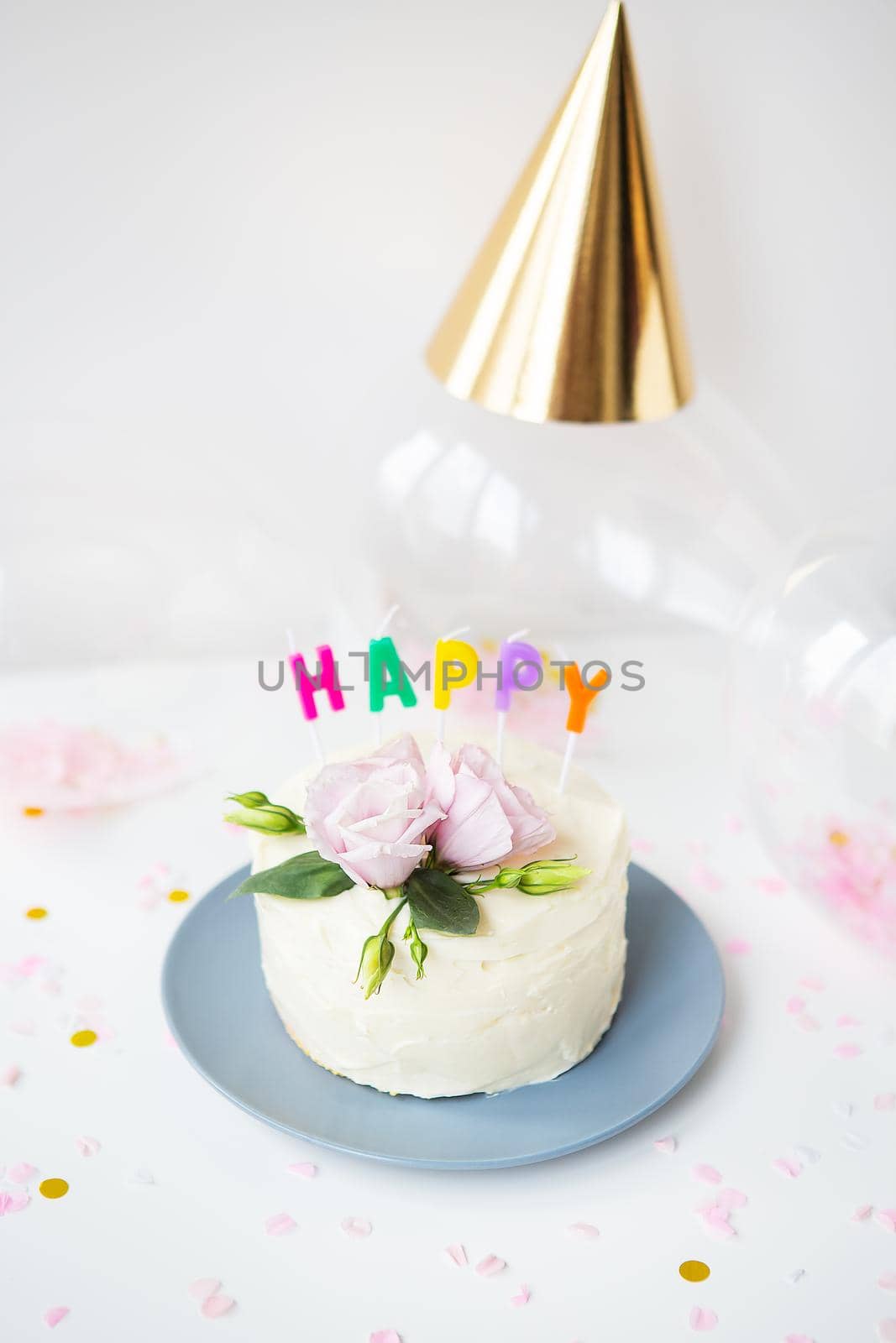 A very beautiful small white cake, decorated with fresh eustoma flowers against the background of sweets and the inscription happy birthday, balloons, hats. Holiday concept
