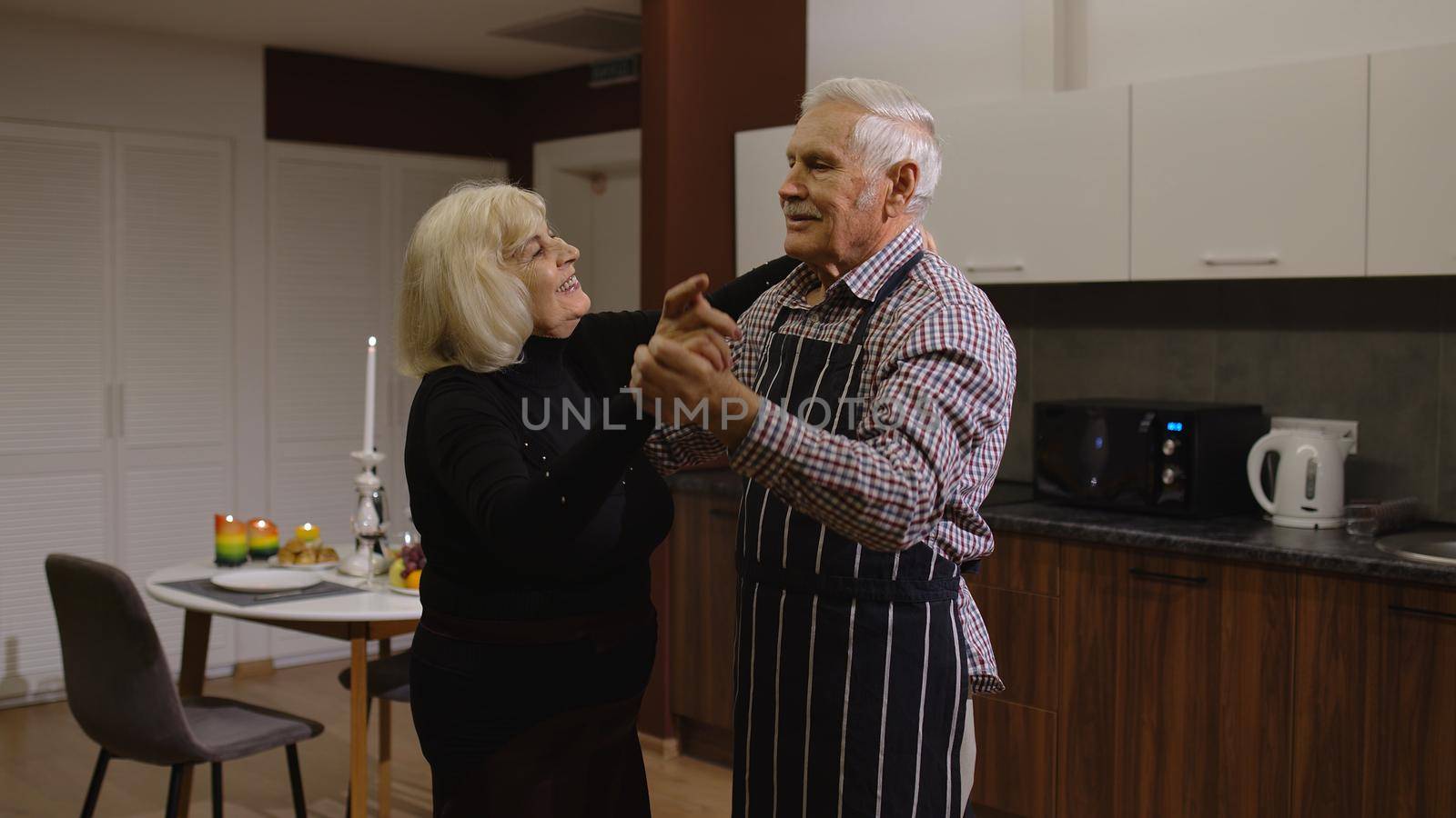 Happy senior couple in love dancing. Romantic evening supper with wine and candles, celebrating anniversary in the kitchen at home. Beautiful romantic elderly grandparents relaxing together enjoy care