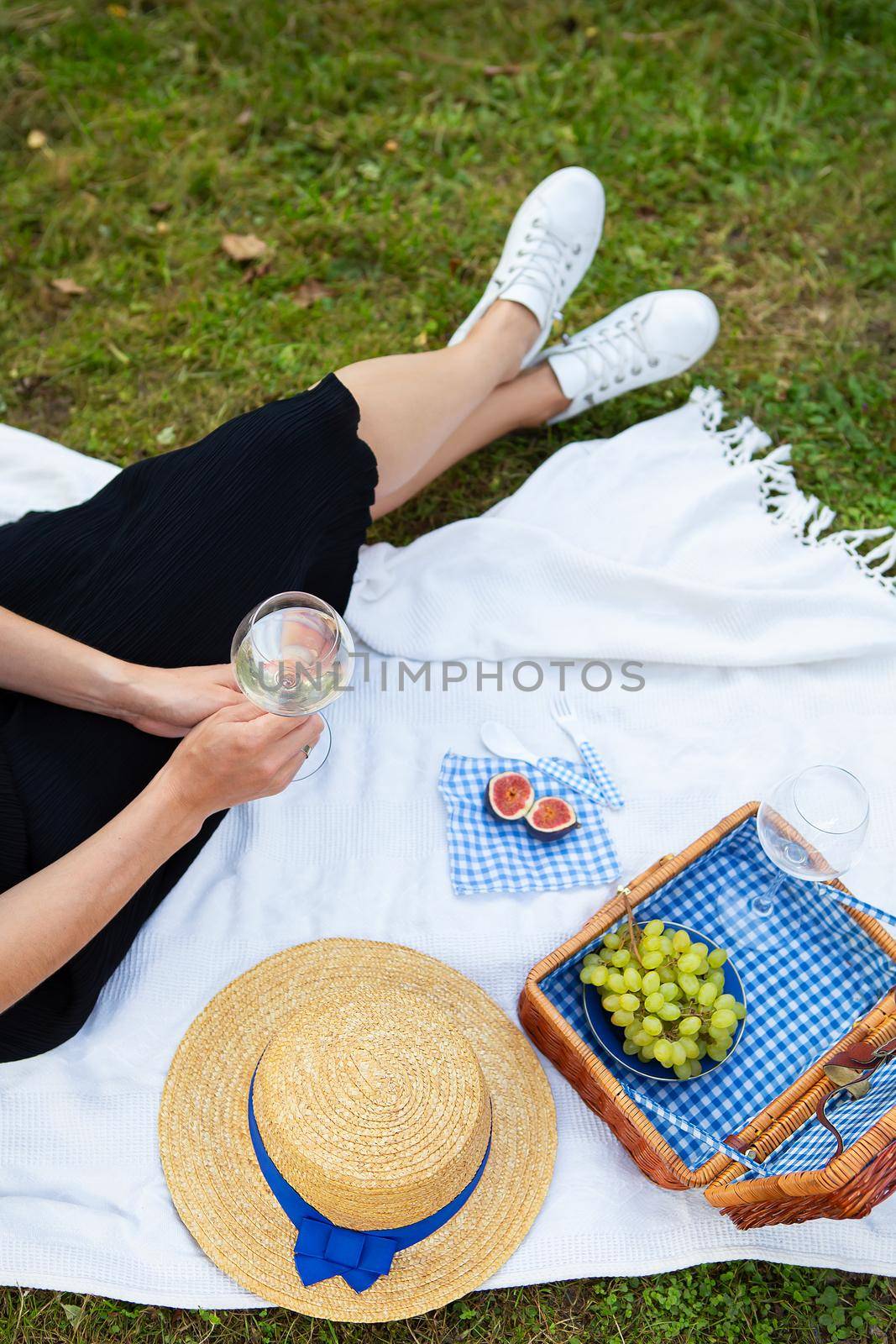 Romantic picnic in the park on the grass, delicious food: basket, wine, grapes, figs, cheese, blue checkered tablecloth, two glasses of wine. The girl is holding a glass of wine in her hands.The concept of outdoor recreation