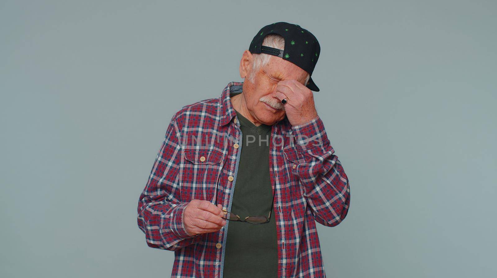 Exhausted tired man 80s in shirt takes off glasses, feels eyes pain, suffering, being overwork burnout stress from long hours working. Mature grandfather isolated alone on gray studio wall background