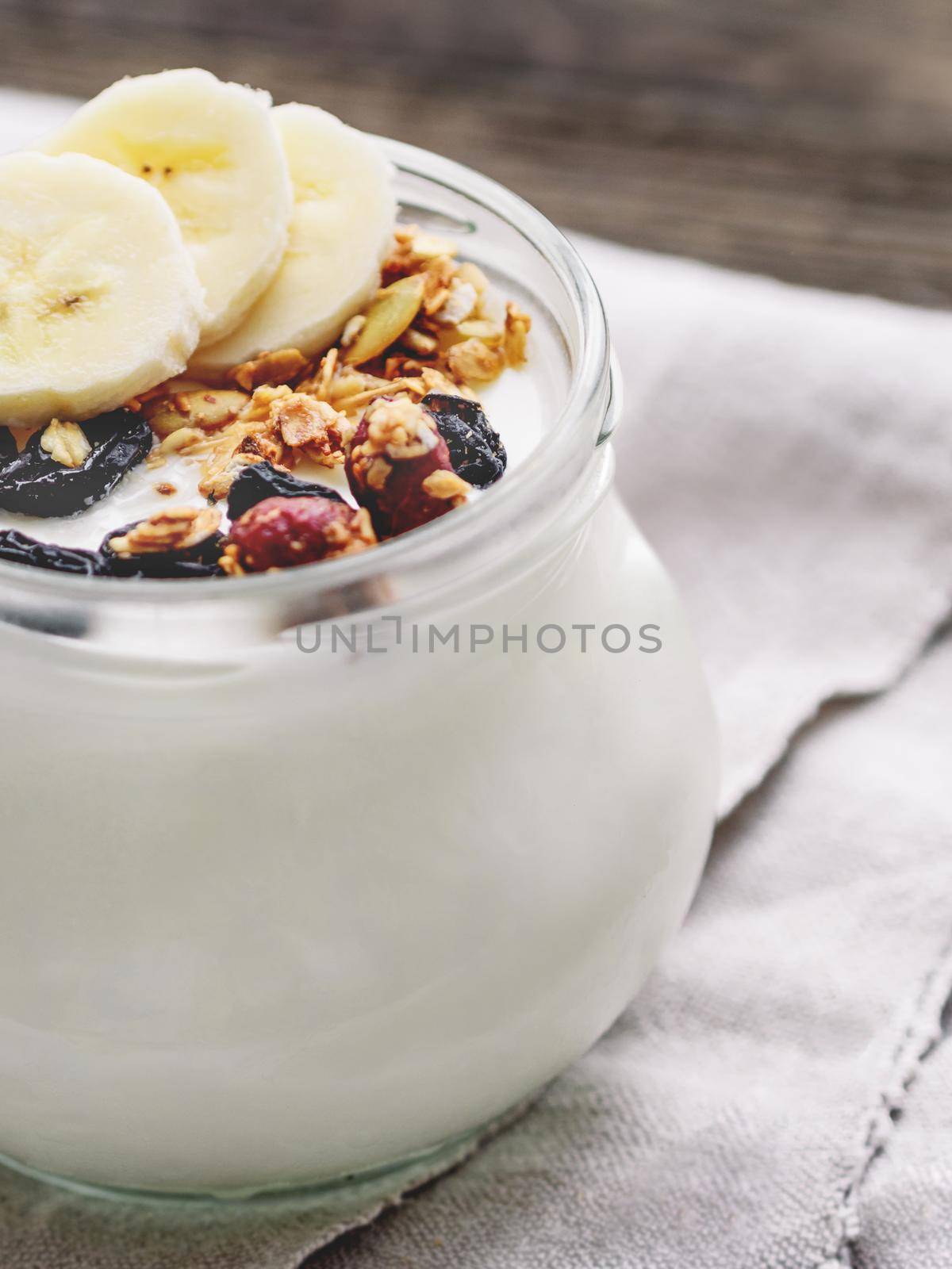 Natural homemade yogurt in a glass jar. Healthy food for breakfast with muesli. Jar with granola and banana slices on linen tablecloth on on wooden table. by aksenovko