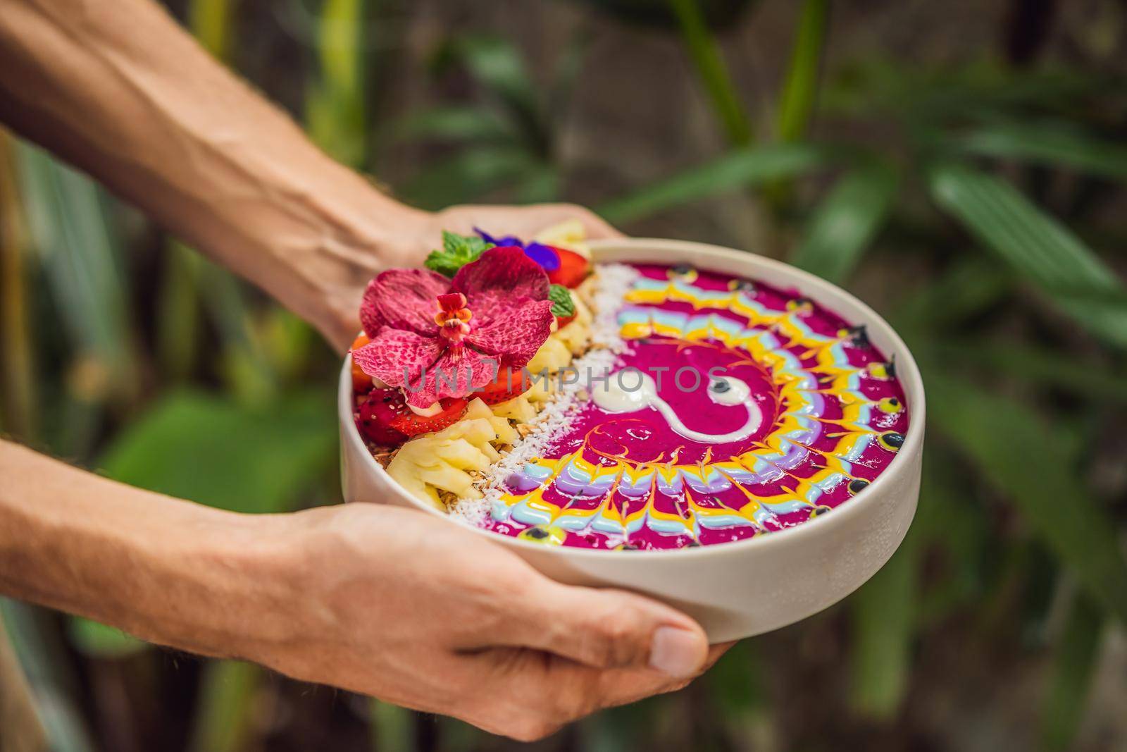 Young man having a mediterranean breakfast, eats Healthy tropical breakfast, smoothie bowl with tropical fruits, decorated with a pattern of colorful yogurt with turmeric and spirulina. It is also decorated with fruits, flowers, chia seeds, coconut, granola, pineapple, mint and strawberries.