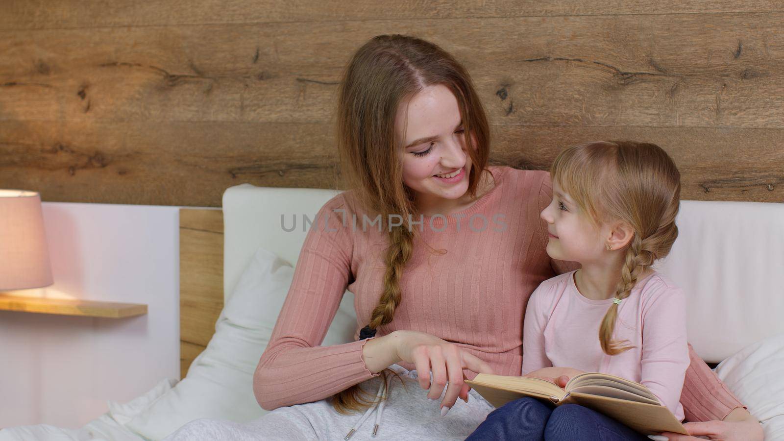 Young mother and teen child daughter lying in bed at home bedroom on duvet blanket and reading book bedtime stories and fairytales together with warm lamp at night. Family bonding, parenthood concept