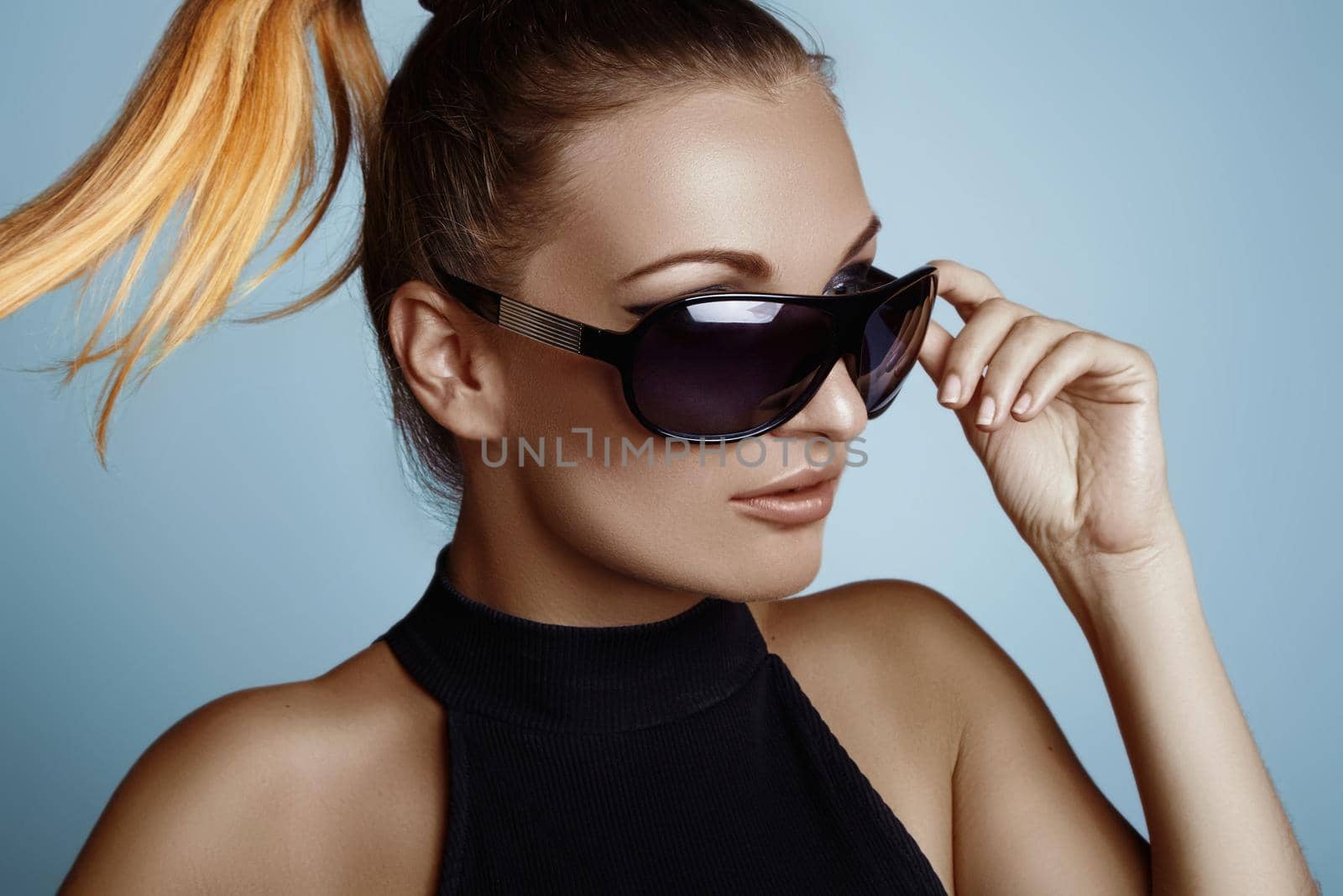 Beautiful young woman with black fashion sunglasses and glamour ponytail hairstyle on blue background. Trendy look for lady. Eye wear style.