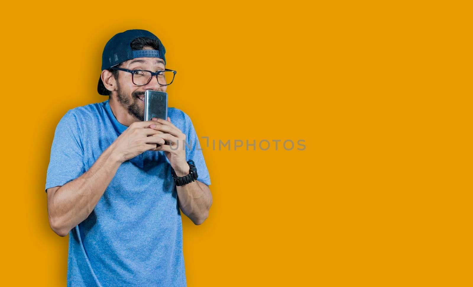 boy in glasses smiling with his cell phone, young man in cap smiling while holding his cell phone on yellow background, boy smiling covering his mouth with cell phone