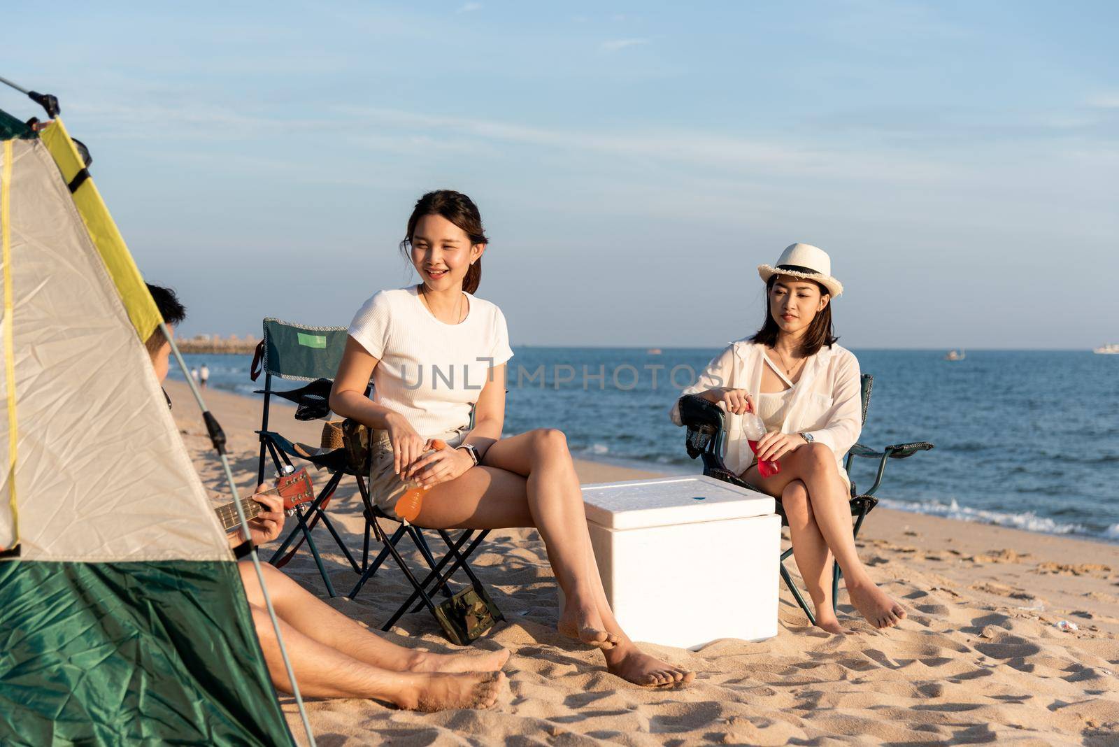 Happy friends with drinks in camp and laughing together in holidays on sand beach near camping tent vacation time at sunset by Sorapop