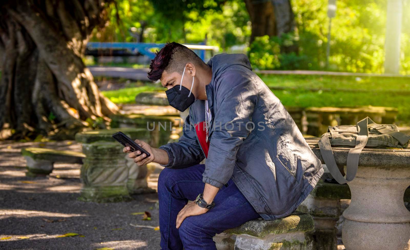 Man with a mask sitting on a bench with his phone, young man with a mask sitting with a cell phone, near a young man with a mask texting