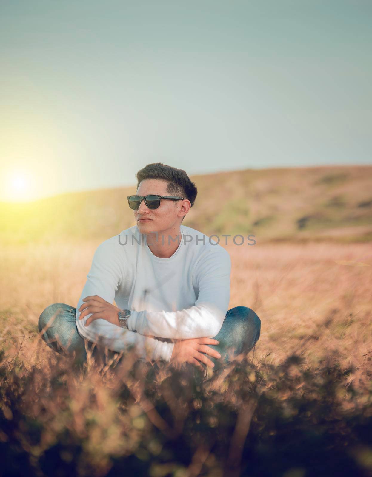 Smiling young man sitting on the grass, smiling young man in eyeglasses sitting on the field, concept of happiness in the field by isaiphoto