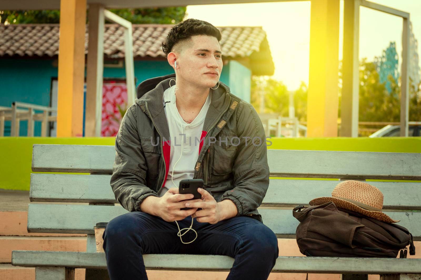 An attractive guy listening to music on a bench, Latin man sitting on a bench listening to music with his phone