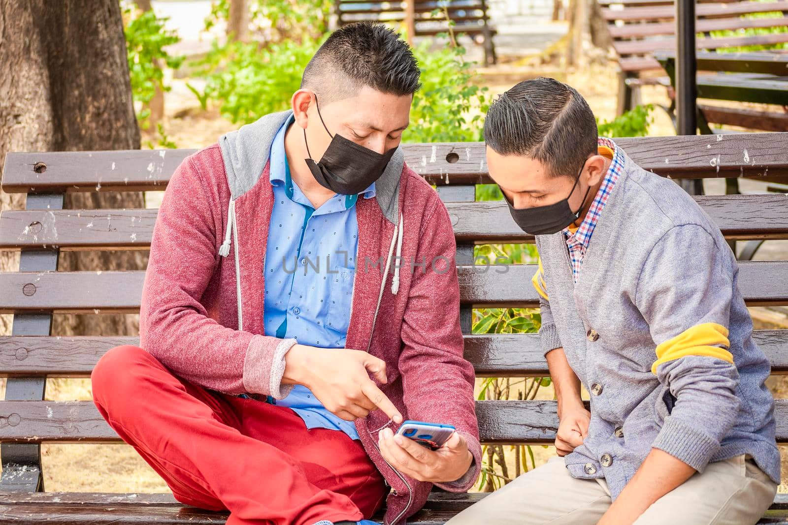 Two men wearing masks checking their cell phones on a bench, a man showing his cell phone to another man