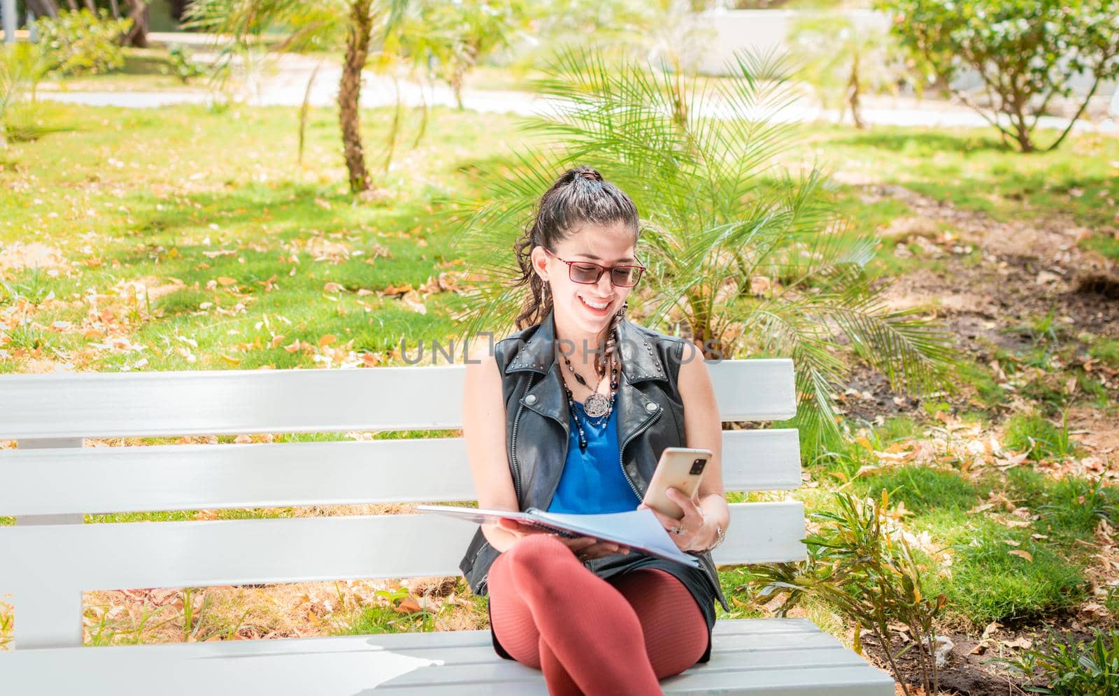 A girl sitting taking notes with a notebook and cell phone, Woman sitting taking notes with her cell phone, Latin girl using her cell phone in a park