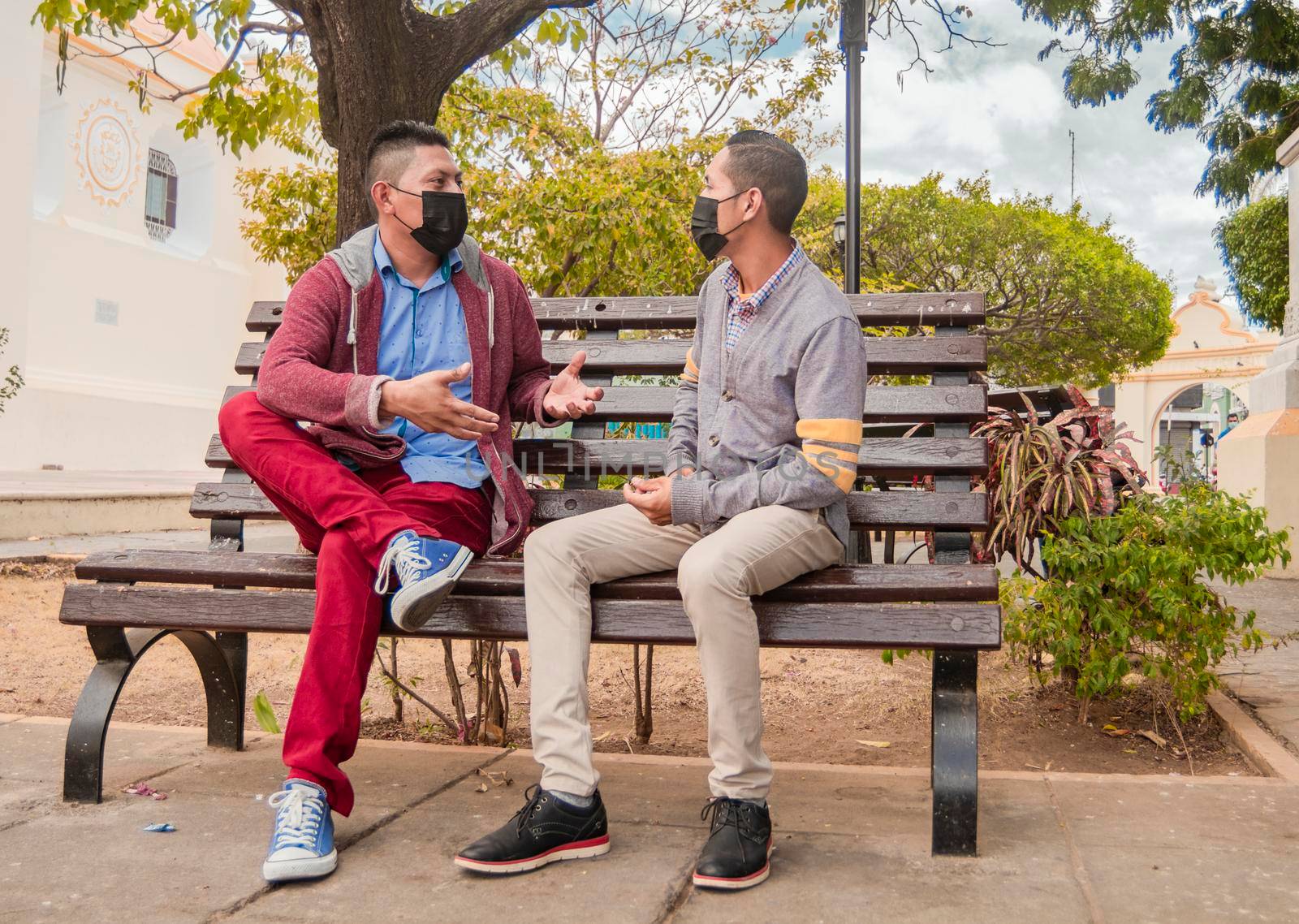 Two men with masks talking on a bench, men with social distance talking on a bench