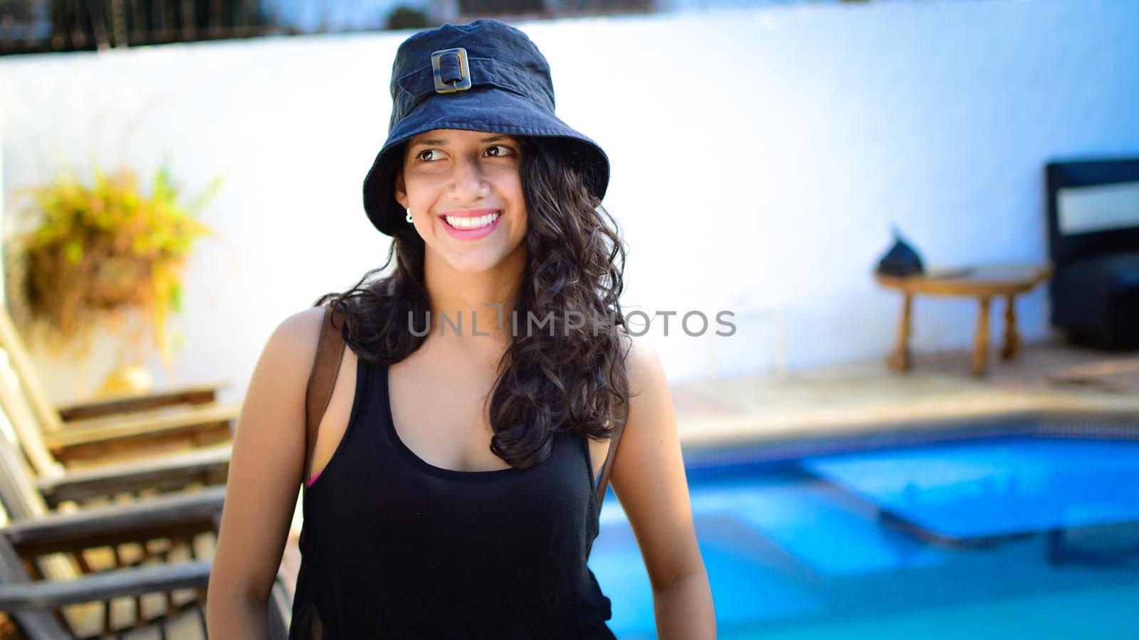 Pretty girl with hat smiling near a swimming pool, latin girl smiling by a swimming pool with muskoka chairs by isaiphoto