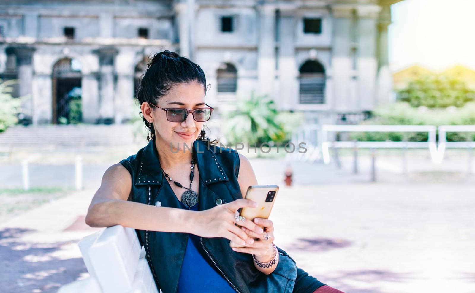 Urban style girl sitting on a bench with her cell phone, An attractive girl sitting on the cell phone smiling, Girl sitting on a bench texting on her cell phone. by isaiphoto