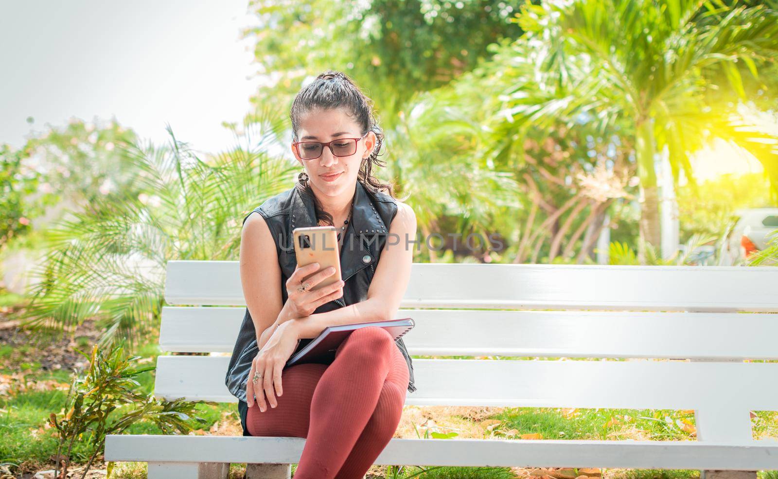 Girl sitting on a bench checking her cell phone, Happy woman sitting in a park texting on her cell phone, Woman sitting on a bench sending a text message