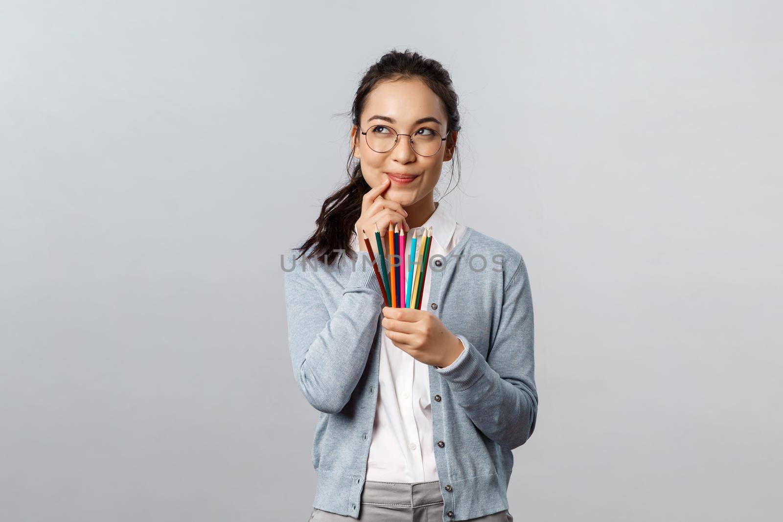 Hobbies, creativity and art concept. Scheming, creative and smart young talented student thinking-up what draw next, holding colored pencils, smirk and look up thoughtful, grey background by Benzoix