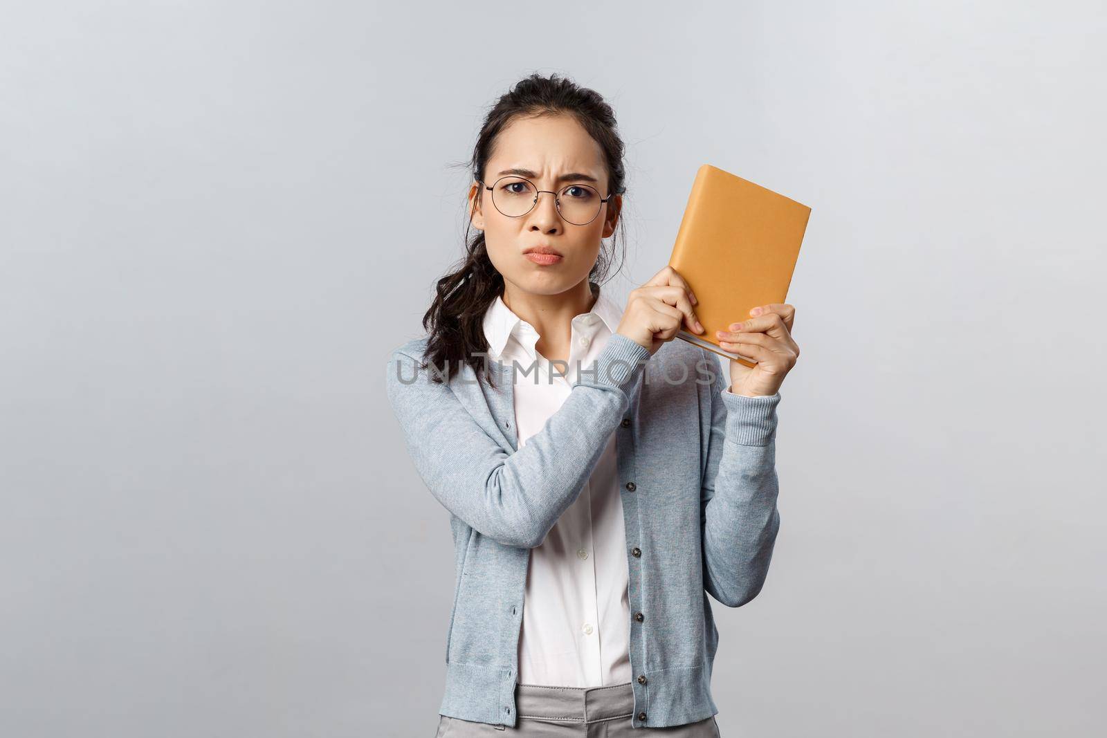 Education, teachers, university and schools concept. Angry displeased, pouting asian girl raise planner want to bit someone with book as hear rude joke, frowning and sulking displeased, threatening.