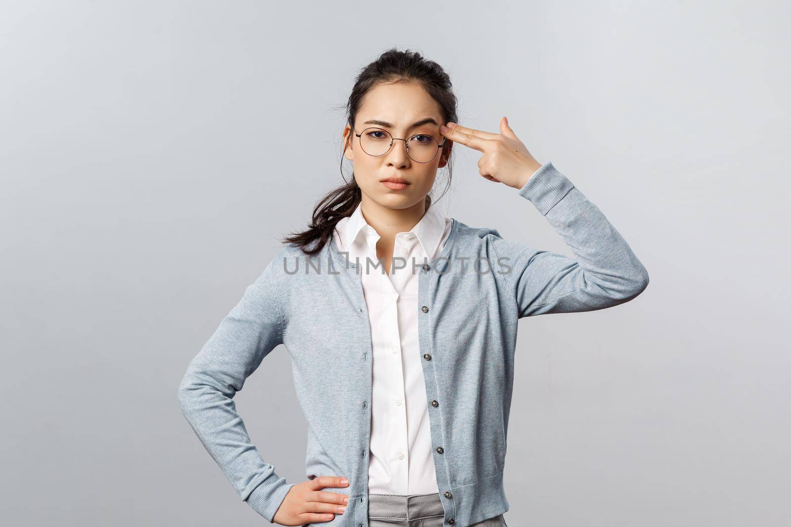 Portrait of sick and tired asian woman hear something stupid, shoot herself with finger gun as a reaction to annoying bothering conversation, staring skeptical camera, standing grey background.