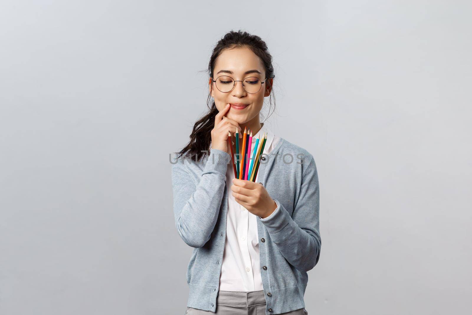 Hobbies, creativity and art concept. Thoughtful young creative female artist touch lip thinking and smiling as look at colored pencils, plan what to draw next for inktober, stand grey background by Benzoix