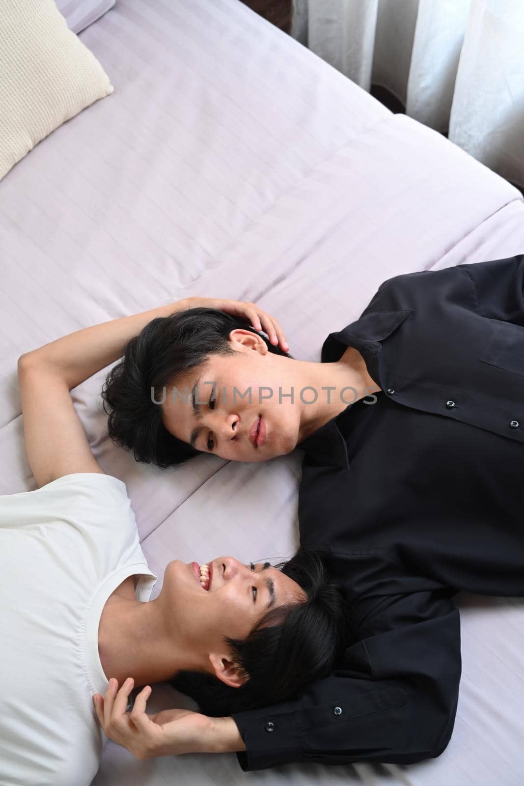Happy young gay couple lying down on bed, spending time together at home. Concept of sexual freedom and equal rights for LGBT community.
