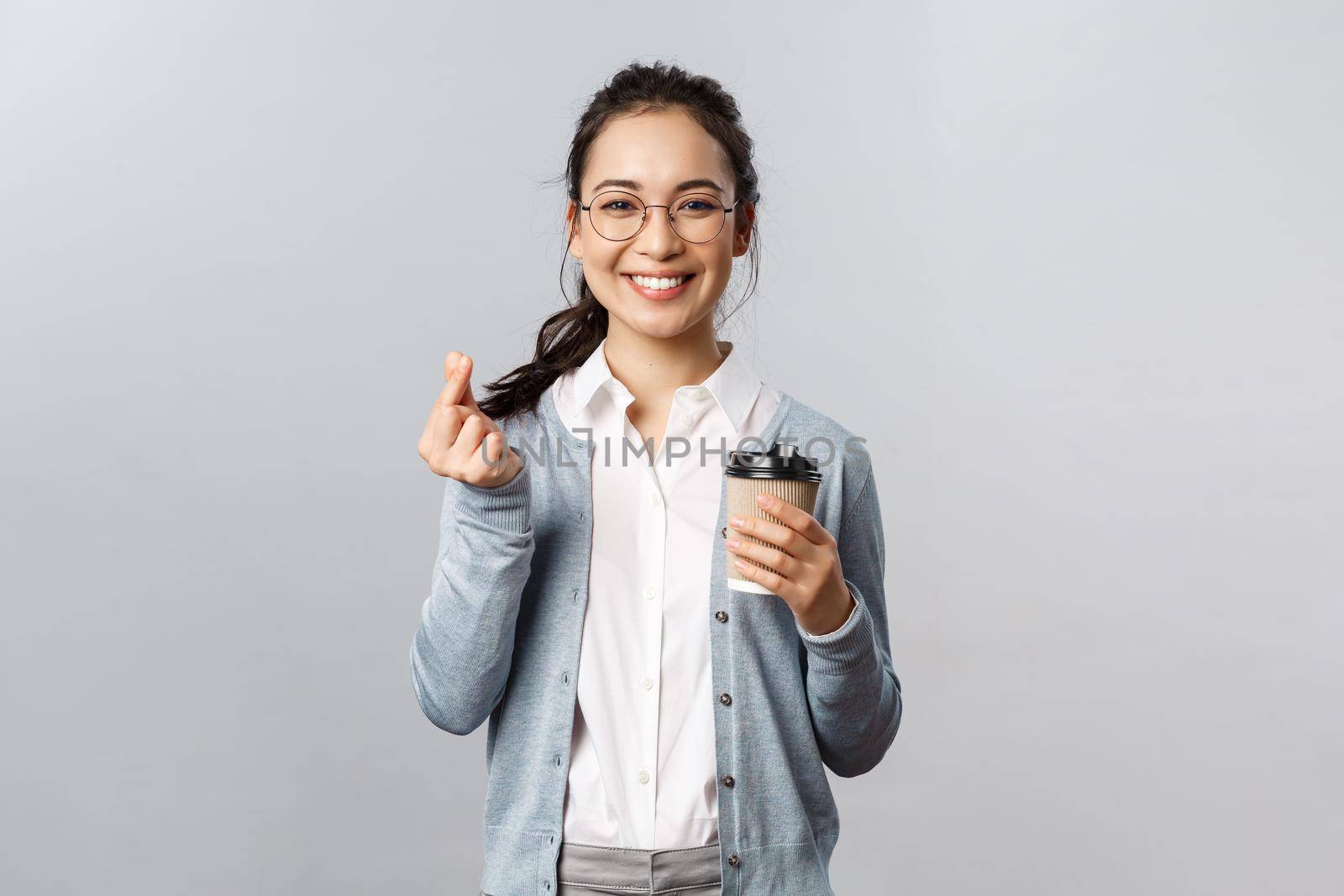 Office lifestyle, business and people concept. Friendly attractive asian female employee, girl showing korean heart with fingers and smiling, drinking take-away coffee, recommend visit cafe.