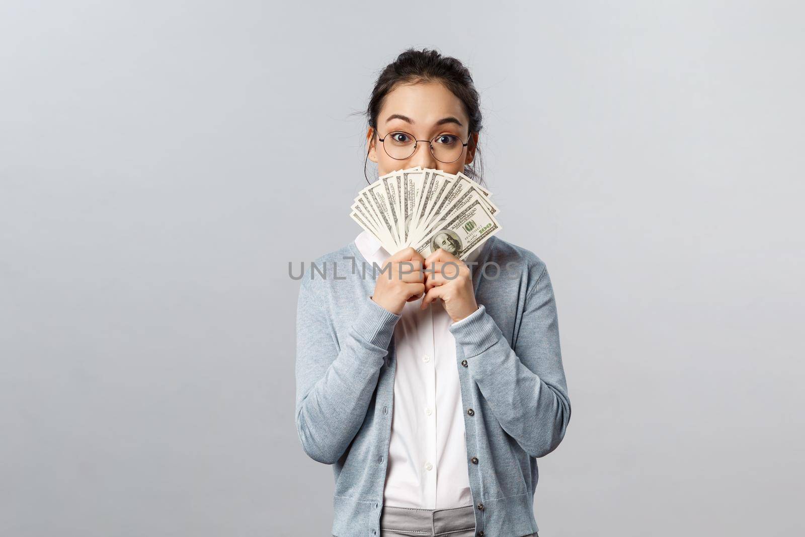 Business, finance and investment concept. Silly happy asian woman in glasses, holding cash of dollars near face, cover big pleased smile as delighted receive award, winning money prize.
