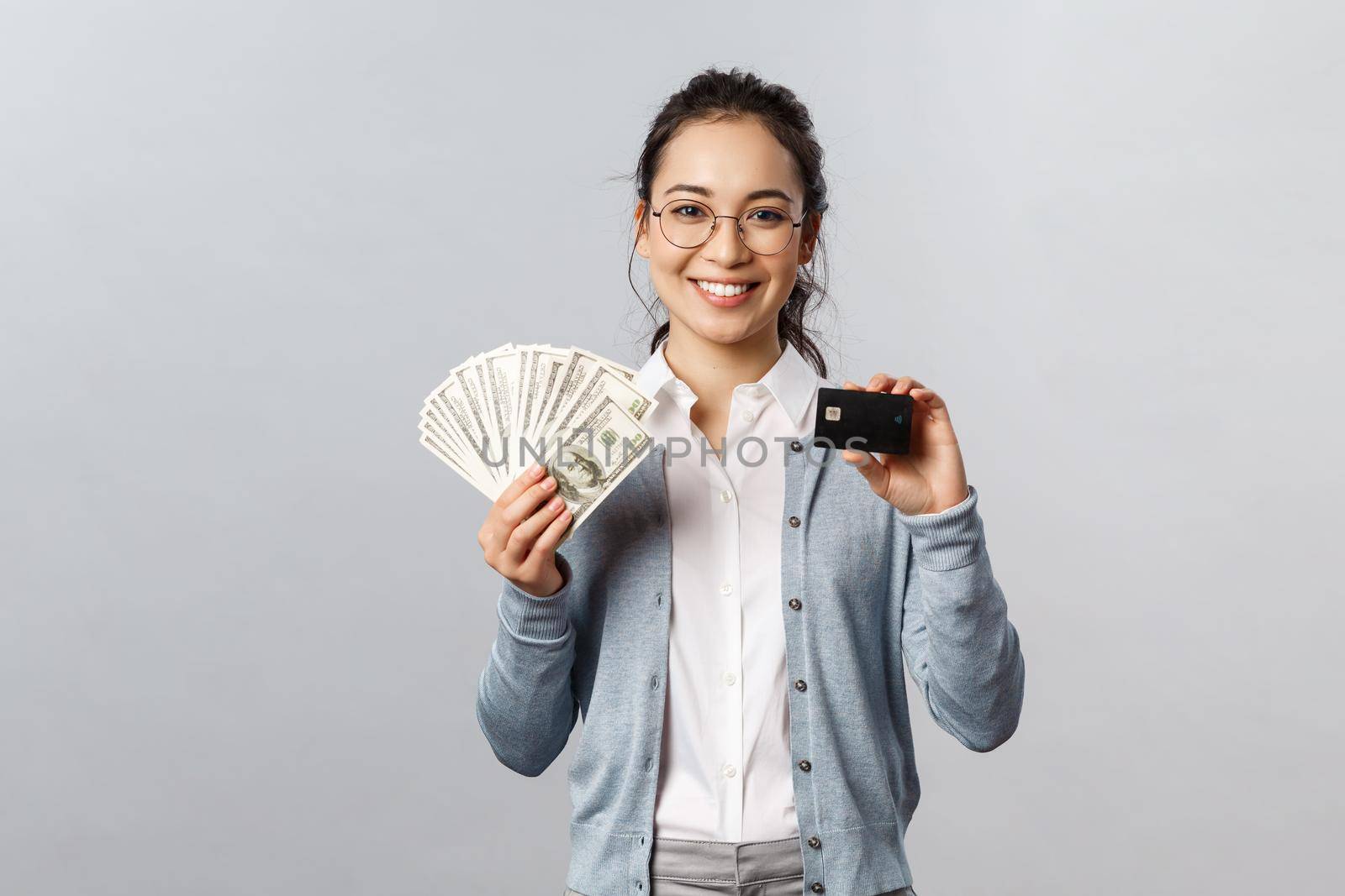 Business, finance and investment concept. Young friendly-looking female employee in glasses, asian woman holding cash and credit card, show different ways of paying online or offline purchase.