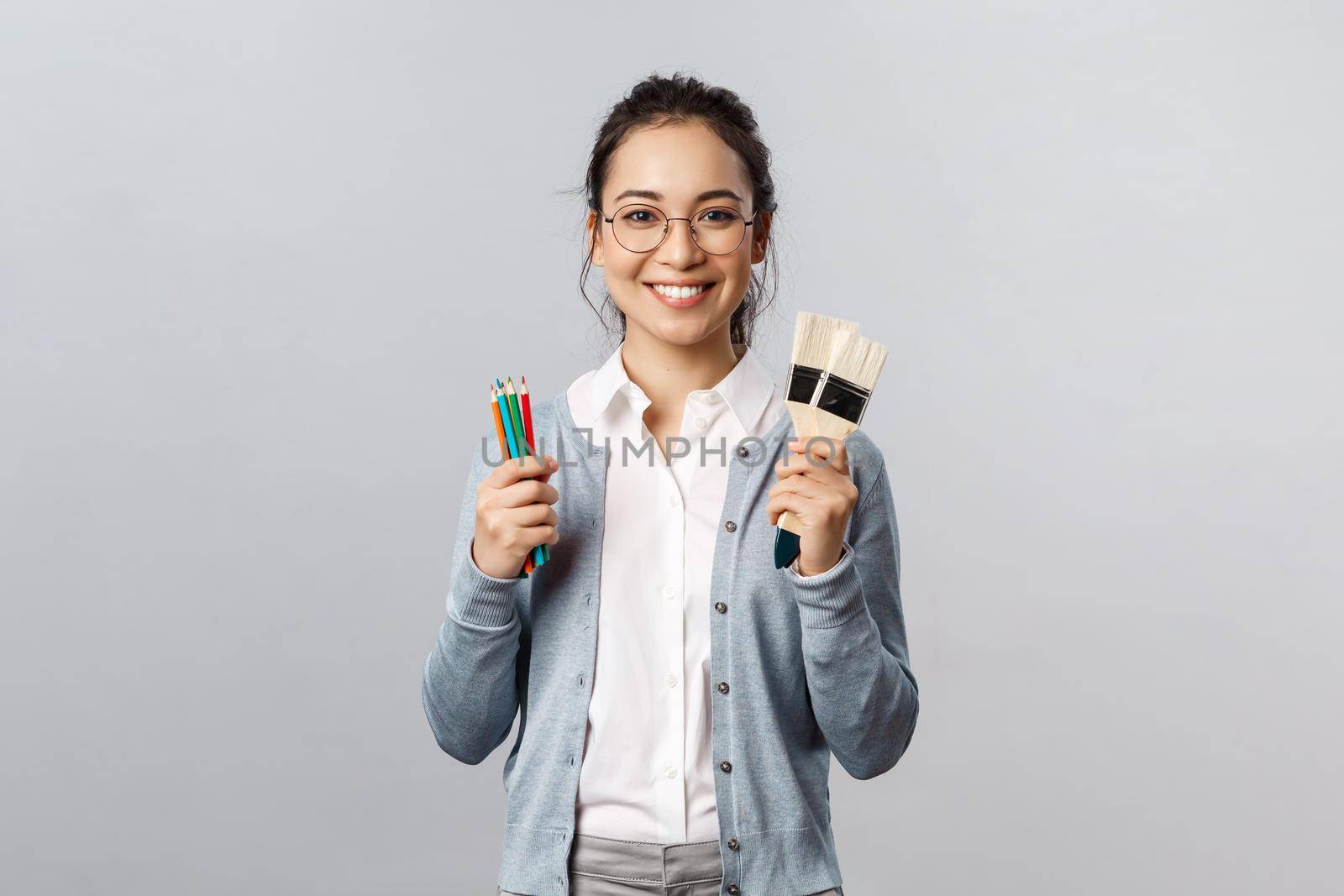 Hobbies, creativity and art concept. Cheerful young asian woman in glasses spend leisure with fun, holding colored pencils and two painting brushes, smiling upbeat, drawing artwork.