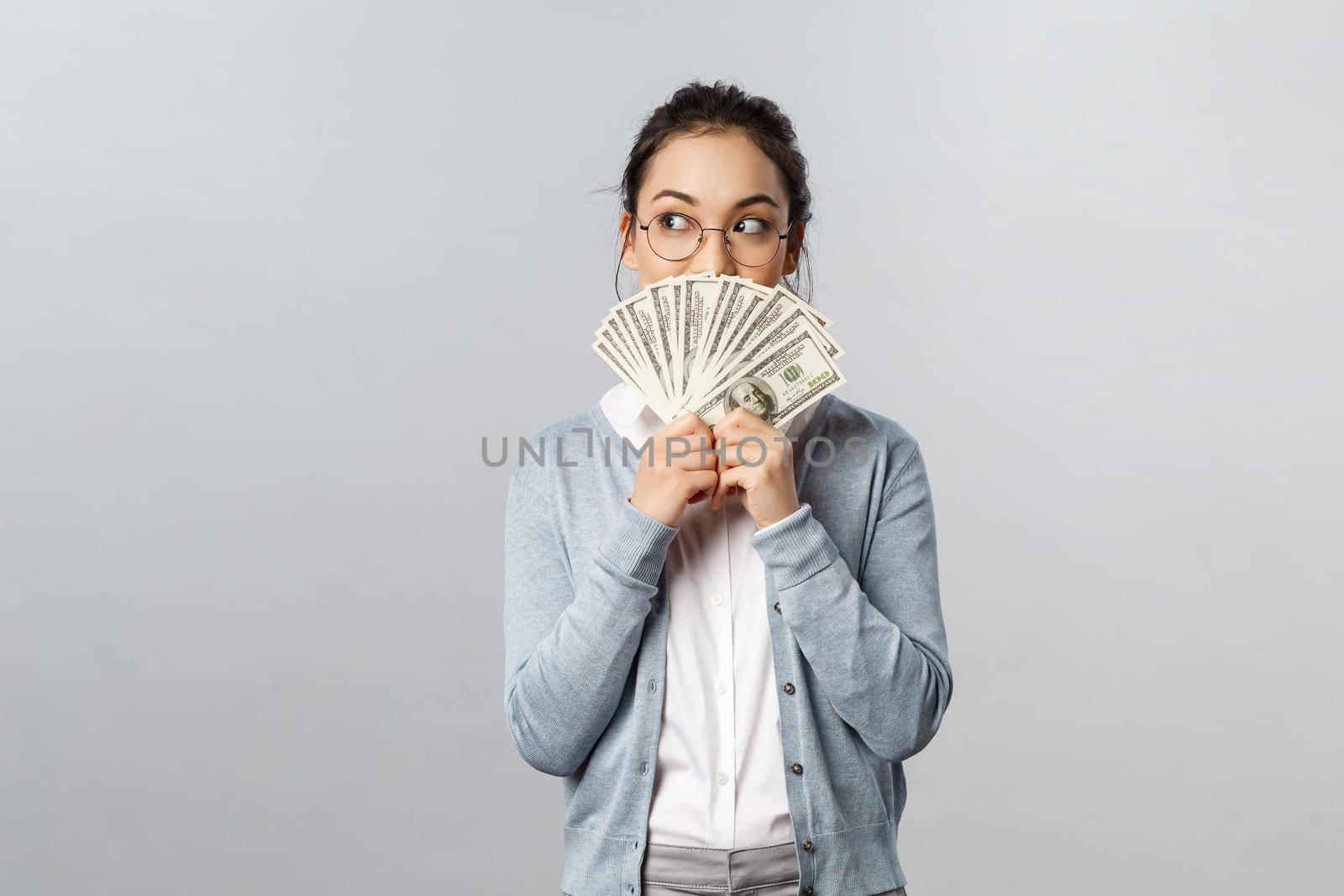 Business, finance and investment concept. Thoughtful excited asian woman looking away pleased and tempting to waste all this money on shopping, look away thinking holding dollars cash.