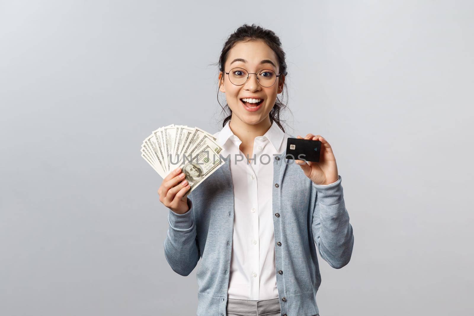 Business, finance and investment concept. Amused happy asian girl decides save for her vacation, place money on deposit, showing cash dollars and credit card, smiling upbeat by Benzoix