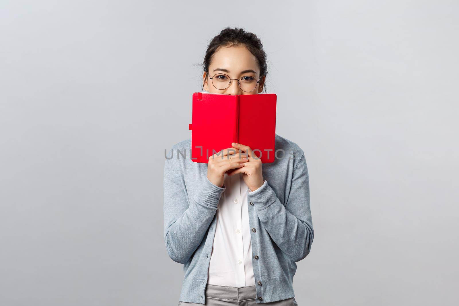 Teaching, education and university lifestyle concept. Silly blushing asian girl hiding her face behind diary, holding notebook or planner, hiding something she wrote from people, smiling with eyes.