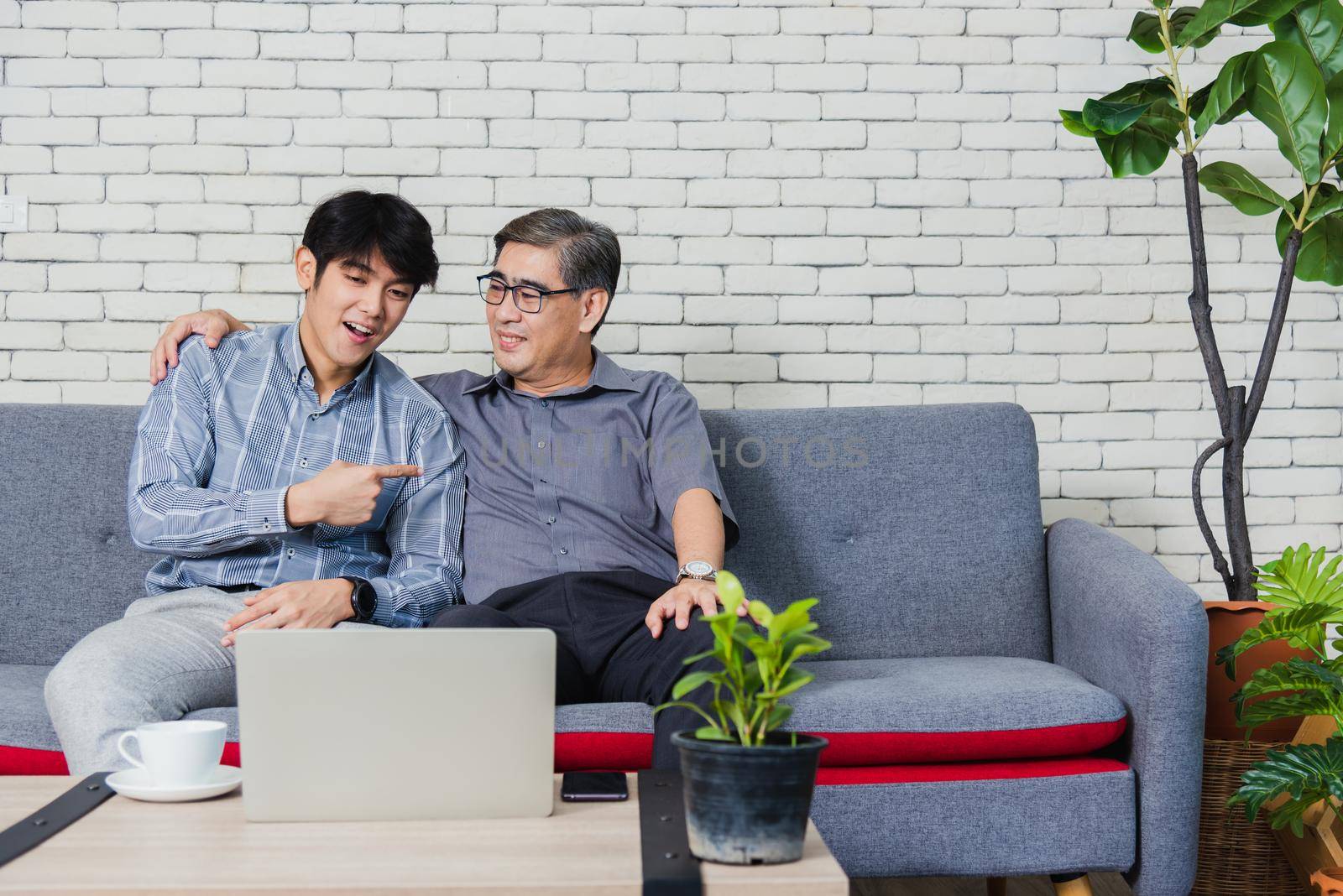 Asian senior businessman with laptop computer discuss together with young team in office. Father man and his son sit on sofa talking chatting on video call conference on laptop in living room at home