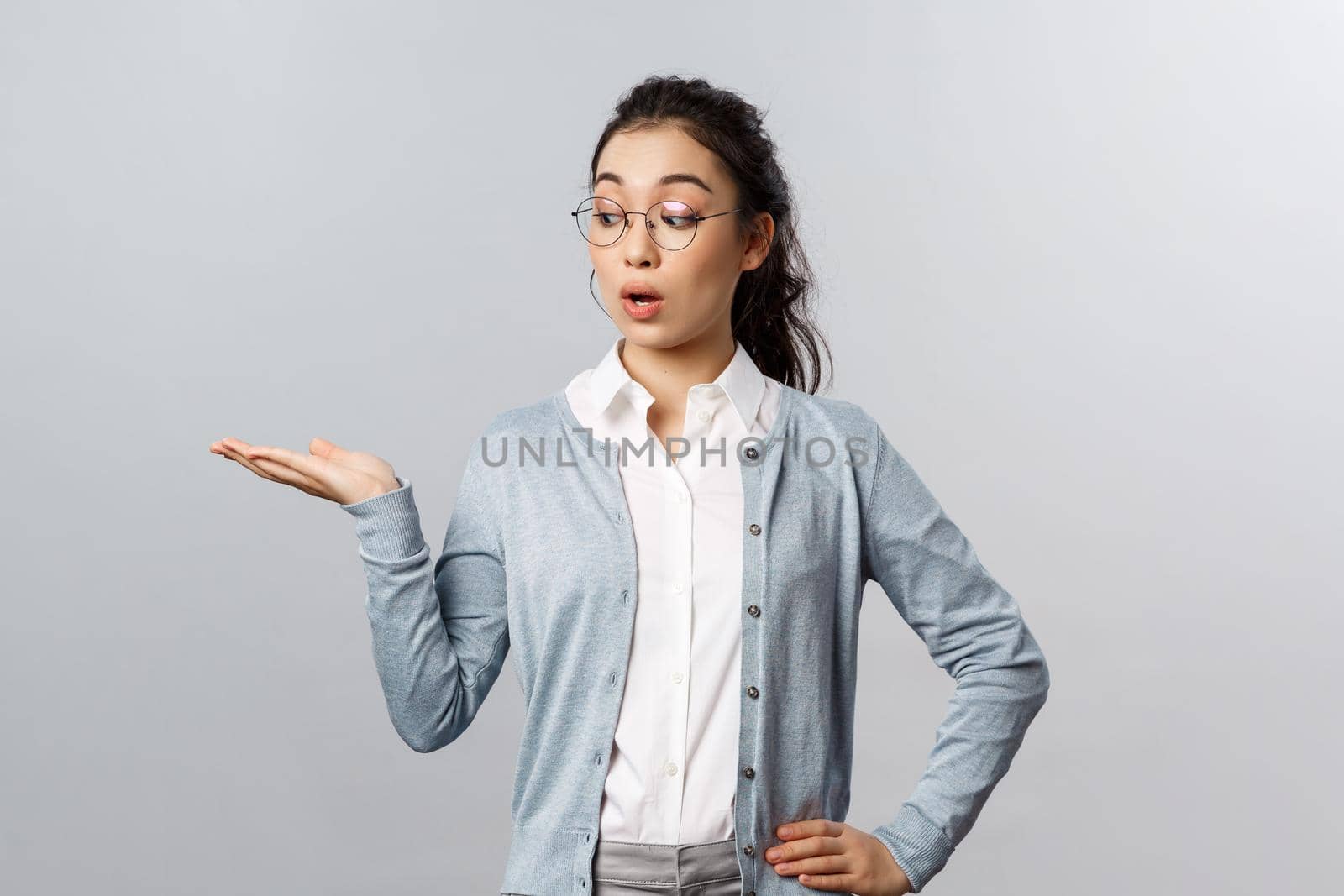 Lifestyle, people and emotions concept. Impressed, excited good-looking asian woman holding promo product on palm and staring at it with interest and curiosity, stand grey background.