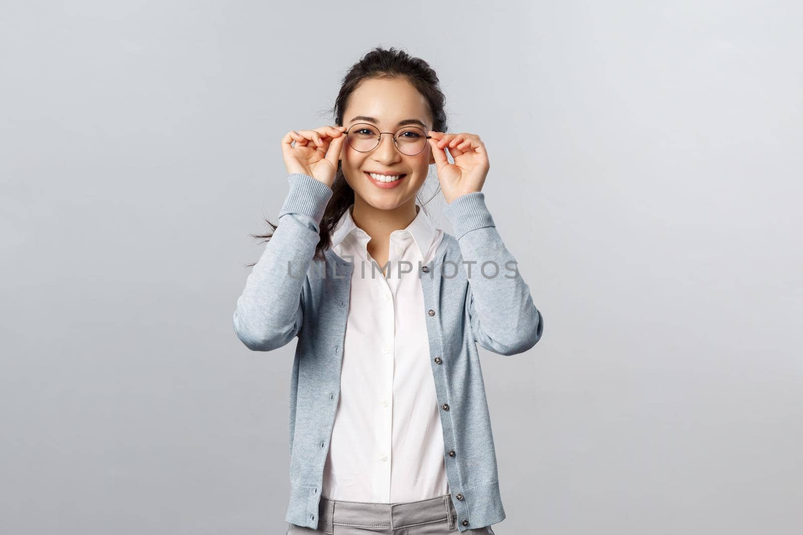 Good-looking young asian girl visit optician store, picking new pair of glasses, put-on eyewear and smiling satisfied, made-up her choice, standing pleased grey background.