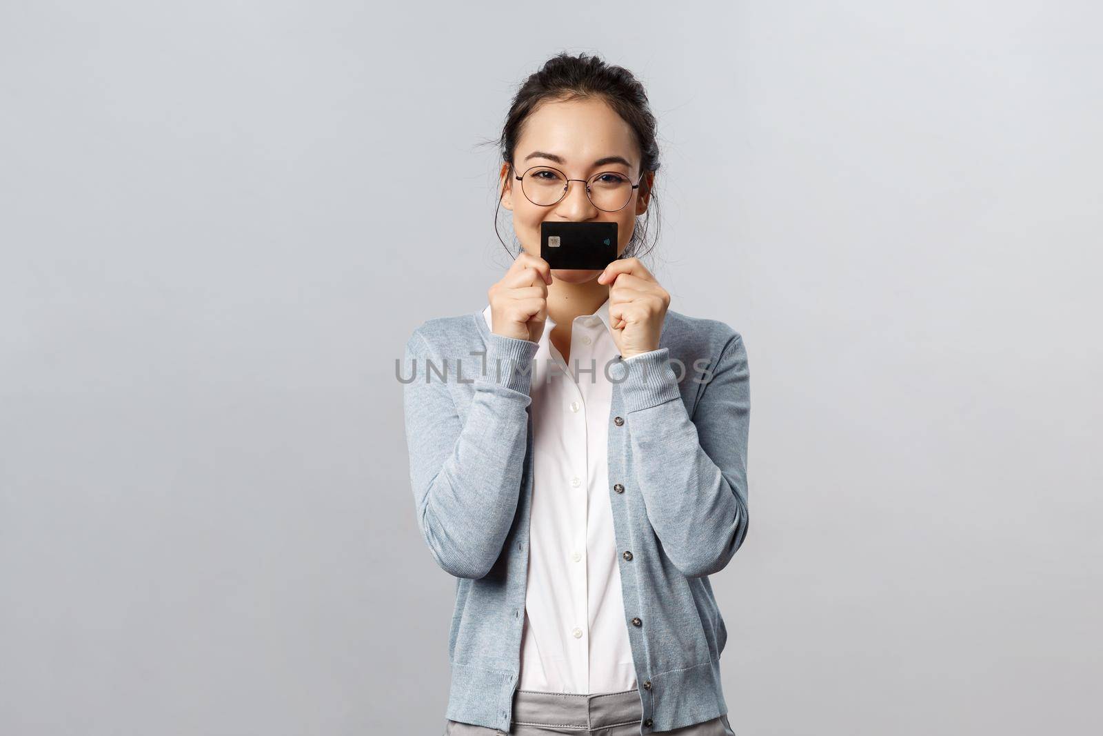 Technology, online purchases and finance concept. Cheerful, smiling asian woman showing her brand new credit card over lips and giggle, cant wait to waste all money on bank account while shopping by Benzoix