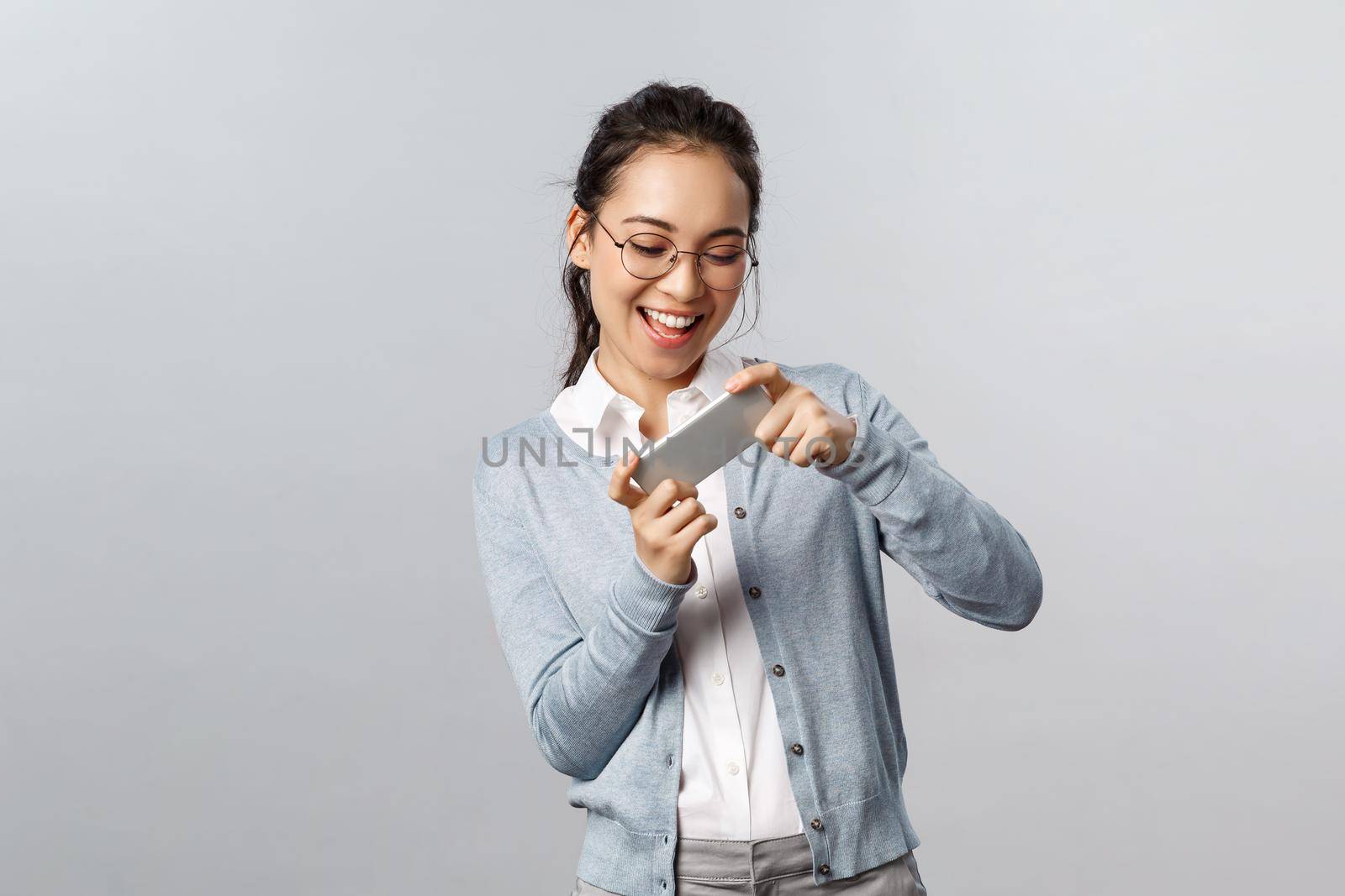 Technology, online and mobile lifestyle concept. Enthusiastic, happy smiling asian woman holding smartphone both hands and tilt phone to pass level, playing racing game amused.