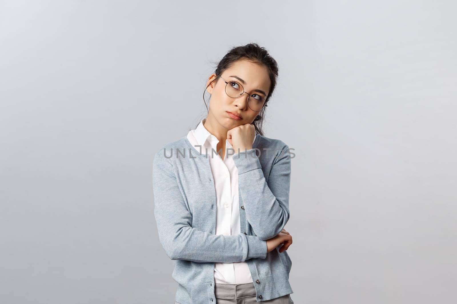 Portrait of bored and tired, thoughtful asian female employer dreaming about home, want go back early from office, looking up dreamy, thinking making plan, use imagination, grey background.