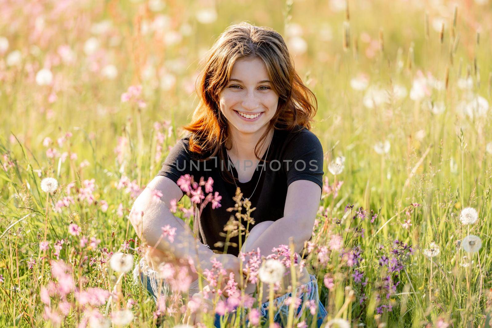 Pretty girl teenager sitting in field with dandelions and flowers, looking at camera and smiling outdoors. Beautiful young female person at nature in sunny day portrait